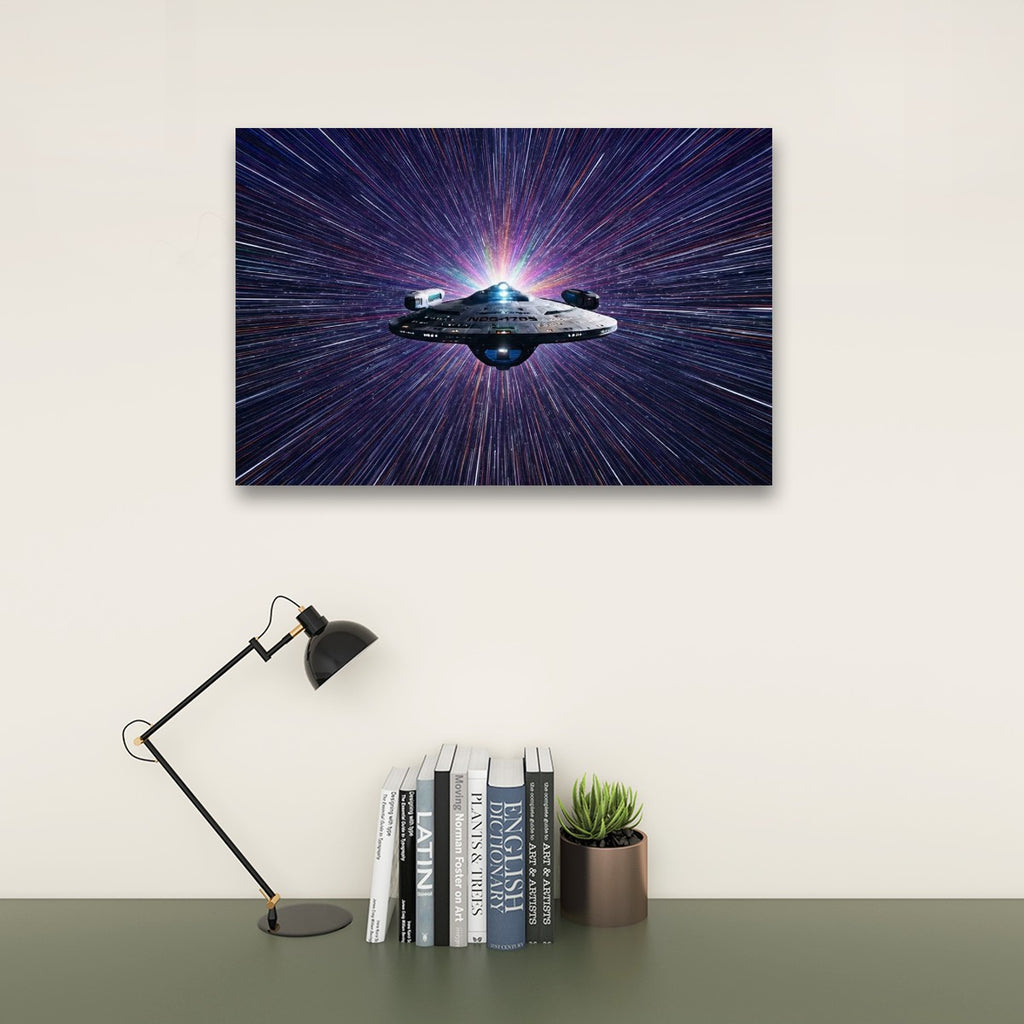 A rectangular wall art print showing a spaceship against a starburst space background, displayed above a desk with books, a small plant, and a black desk lamp in a styled interior setting.