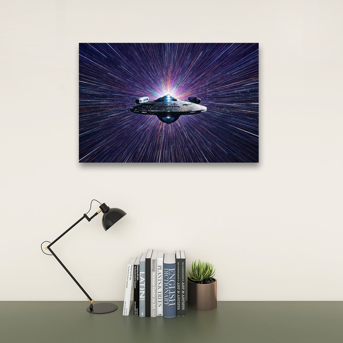 A rectangular wall art print showing a spaceship against a starburst space background, displayed above a desk with books, a small plant, and a black desk lamp in a styled interior setting.