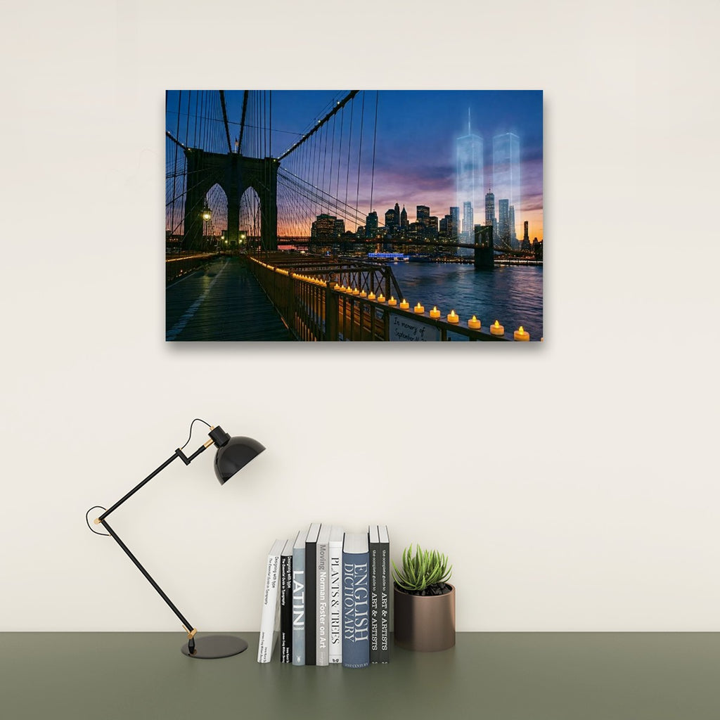 A rectangular wall art print showing a Brooklyn Bridge cityscape at dusk with the Twin Towers ghosted in the skyline, displayed above a desk with a black lamp, books, and a small succulent in a room setting.