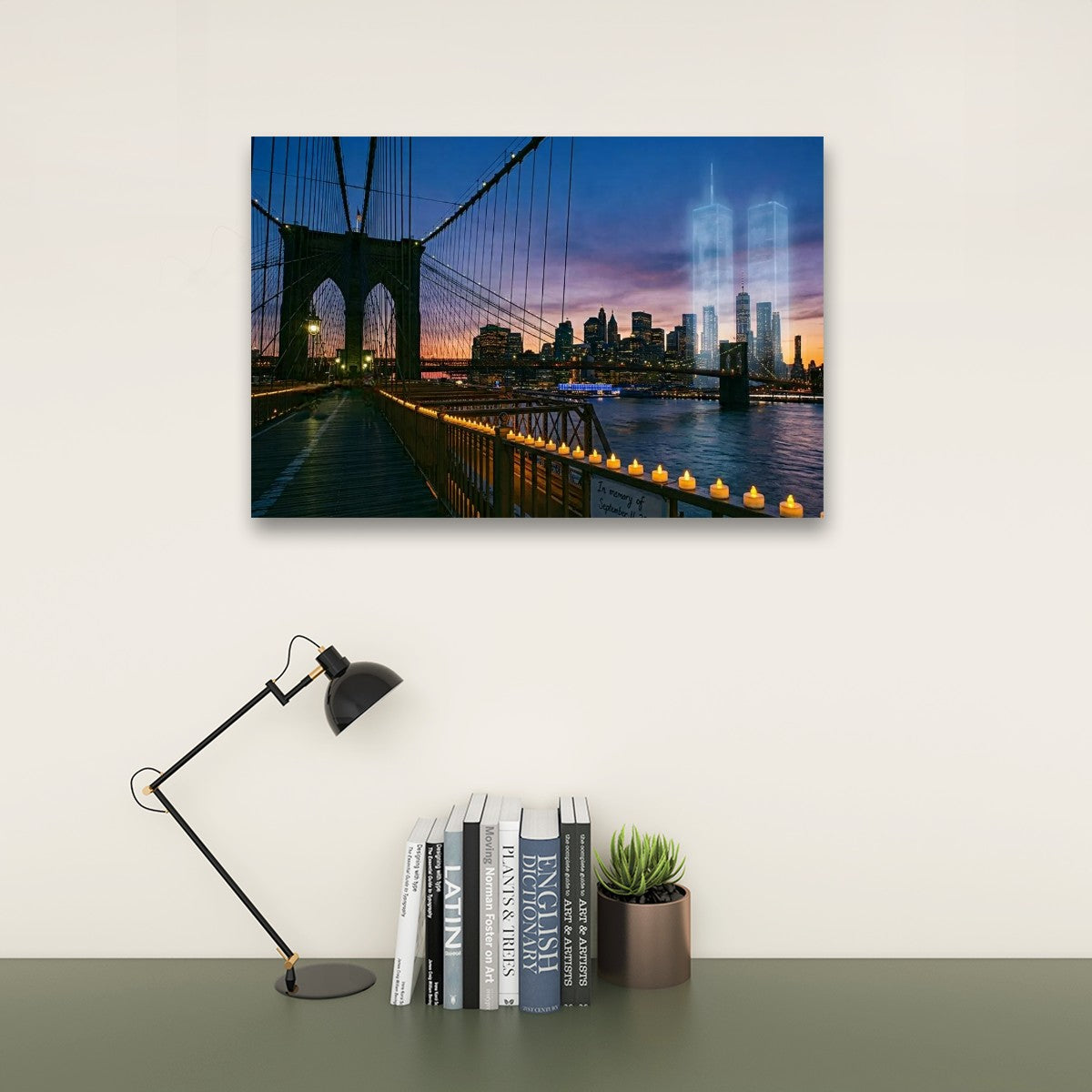 A rectangular wall art print showing a Brooklyn Bridge cityscape at dusk with the Twin Towers ghosted in the skyline, displayed above a desk with a black lamp, books, and a small succulent in a room setting.