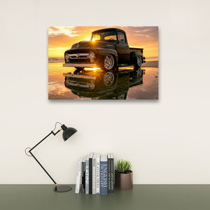 A wall-mounted rectangular art print showing a black  1956 Ford  F-100 vintage pickup truck reflected on wet sand at sunset, displayed above a desk with a black lamp, books, and a small succulent.