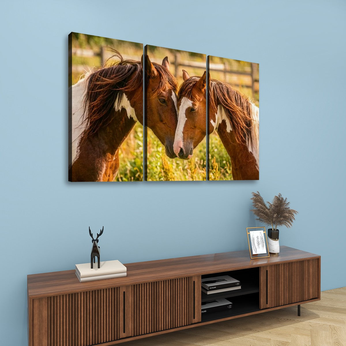 A three-panel canvas wall art set depicting two brown horses with white markings, displayed on a light blue wall above a wooden console table with decorative items.