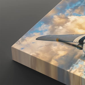 A close-up of a rectangular canvas print showing a printed scene of a propeller airplane wing flying through blue sky with large cream and gold clouds. The canvas is angled to show its side edge against a dark gray background.