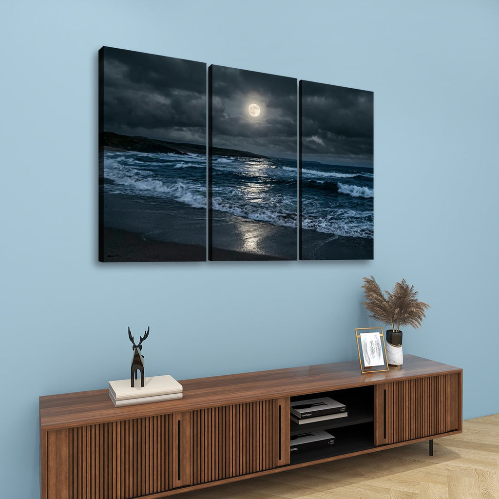 A three-panel canvas wall art set depicting a dramatic moonlit ocean scene with dark waves, a glowing full moon, and a sandy shore, displayed above a wooden console against a light blue wall.