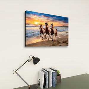 A rectangular wall art print showing three horses running on a beach at sunset, displayed above a desk with a black lamp and books against a light beige wall.