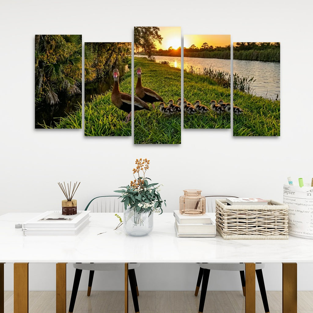 A five-panel canvas wall art set depicting a family of Black-bellied Whistling-Ducks and ducklings by a lake at sunset, displayed on a white wall above a styled dining table with books, a vase, and a basket.