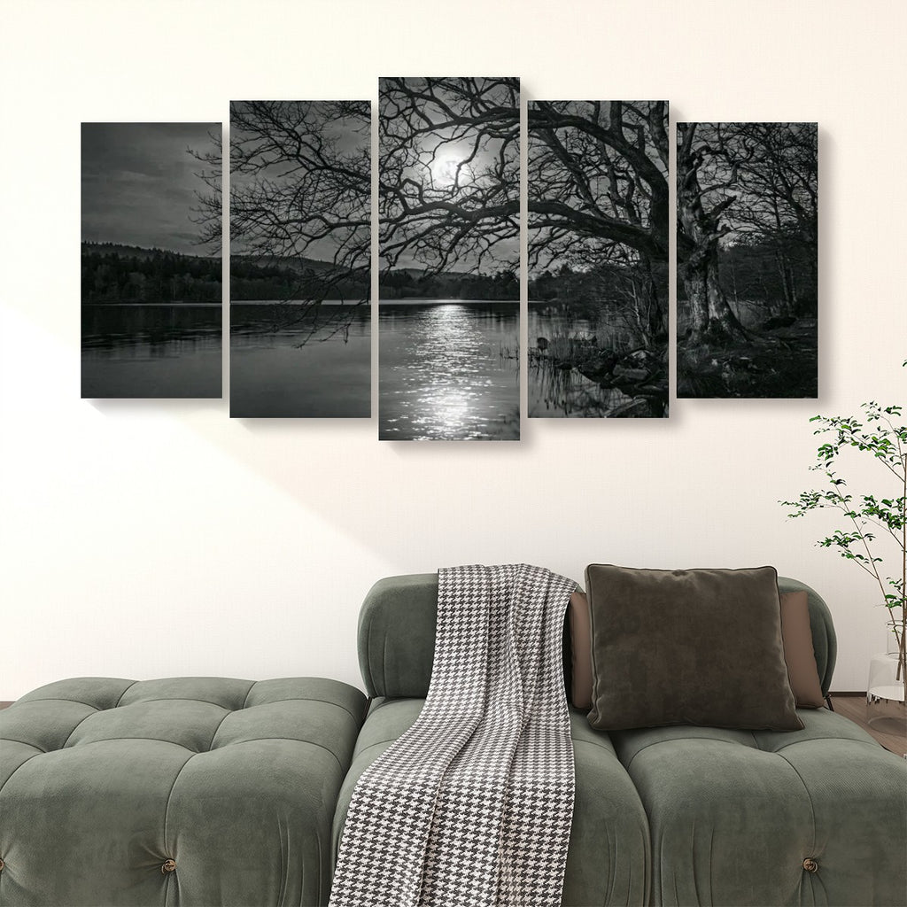 A five-panel black and white canvas wall art set depicting a moonlit lake scene with bare trees, shown hanging above a green sofa in a living room setting.