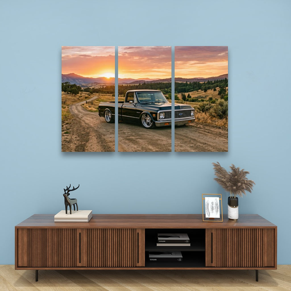 A three-panel canvas wall art print depicting a vintage pickup truck on a dirt road at sunset, displayed on a light blue wall above a wooden console table with decorative items.