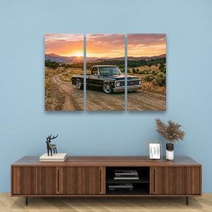 A three-panel canvas wall art print depicting a vintage pickup truck on a dirt road at sunset, displayed on a light blue wall above a wooden console table with decorative items.