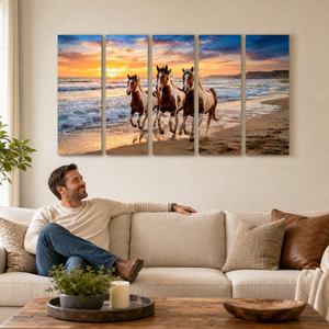 A man sits on a beige sofa in a living room, looking at a five-panel canvas wall art print depicting three horses running on a beach at sunset.