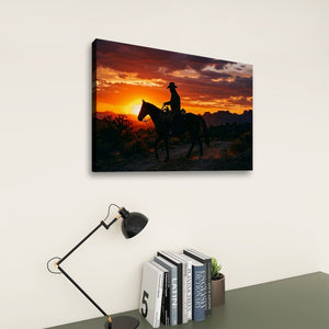 A rectangular wall art print showing a cowboy riding a horse at sunset, displayed on a light wall above a desk with a black lamp and books.