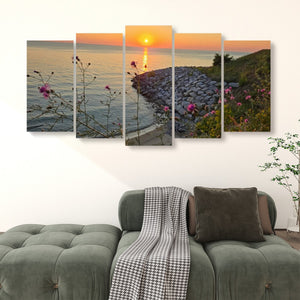 A five-panel canvas wall art set depicting a scenic sunset over a rocky shoreline with pink flowers, displayed above a green sofa in a living room setting.