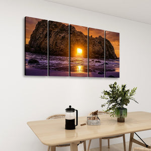 A five-panel canvas wall art set depicting a dramatic rocky coastline at sunset, displayed on a white wall above a light wood dining table with chairs, a coffee press, and a potted plant.