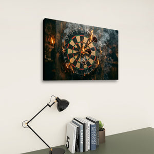 A rectangular wall art print showing a burning dartboard with smoke and flames, displayed above a desk with a black lamp and books against a light beige wall.