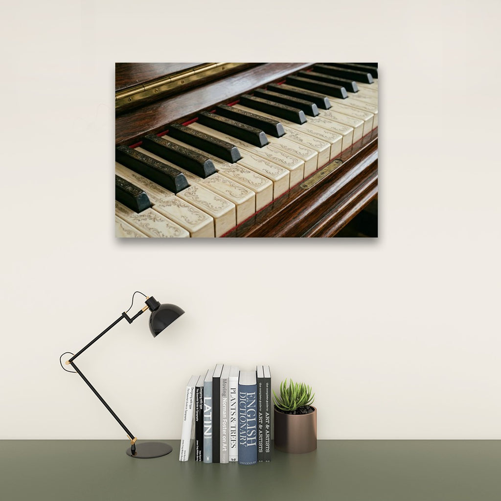 A rectangular wall art print showing a close-up photograph of piano keys and wood casing, displayed above a desk with a black desk lamp, a row of books, and a small potted succulent against a light beige wall.