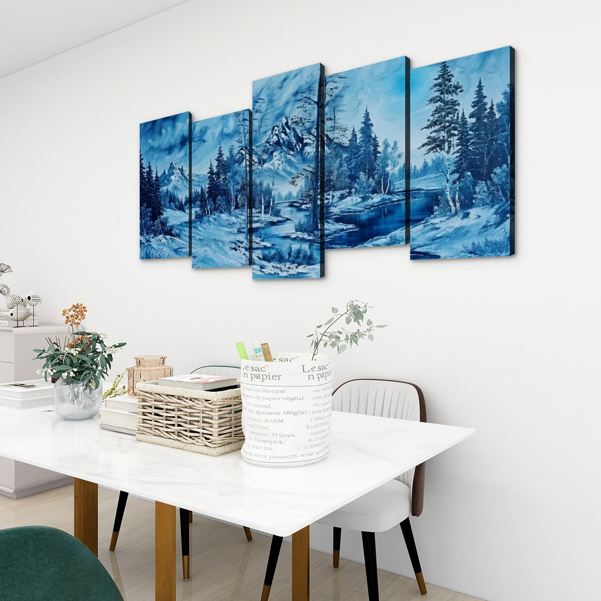 A five-panel canvas wall art set depicting a snowy mountain landscape with pine trees and blue water, displayed above a white dining table in a modern room setting.