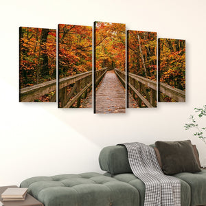 A five-panel canvas wall art set depicting a wooden boardwalk bridge through a vibrant autumn forest with orange and yellow foliage, displayed on a white wall above a green sofa.