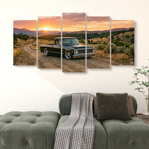 A five-panel canvas wall art print depicting a black vintage 1970 Chevy C10 Fleetside pickup truck driving on a dirt road at sunset, displayed above a green tufted sofa in a living room setting.