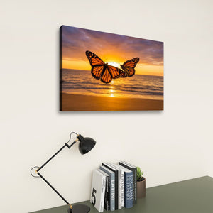 A rectangular wall art print showing two orange monarch butterflies over a beach sunset, displayed on a light wall above a desk with a black lamp and books.