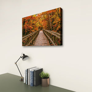 A rectangular wall art print showing a wooden boardwalk bridge surrounded by vivid autumn foliage in orange, yellow, and red, displayed on a light wall above a desk with books, a lamp, and a small plant.