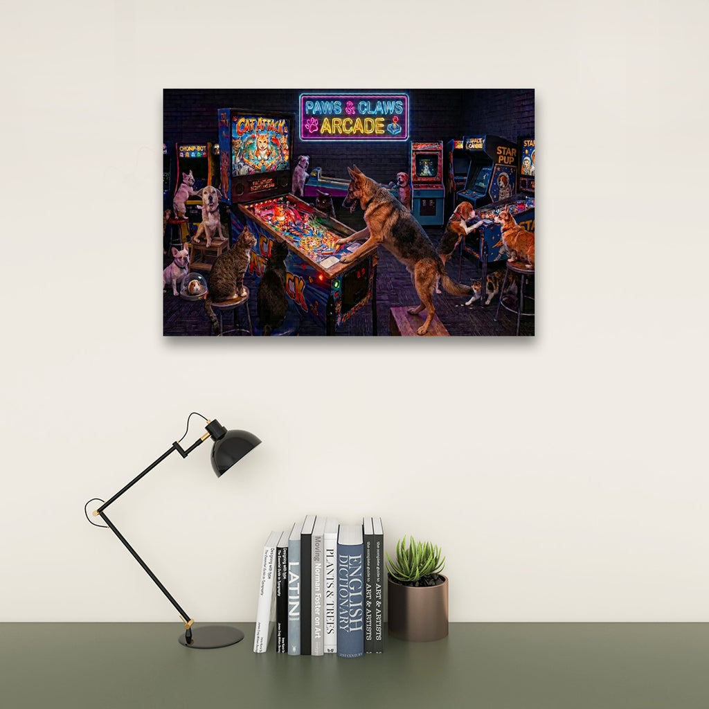 A framed wall art print depicting a retro arcade scene with dogs playing pinball machines, displayed above a desk with a black lamp, books, and a small potted succulent against a light beige wall.
