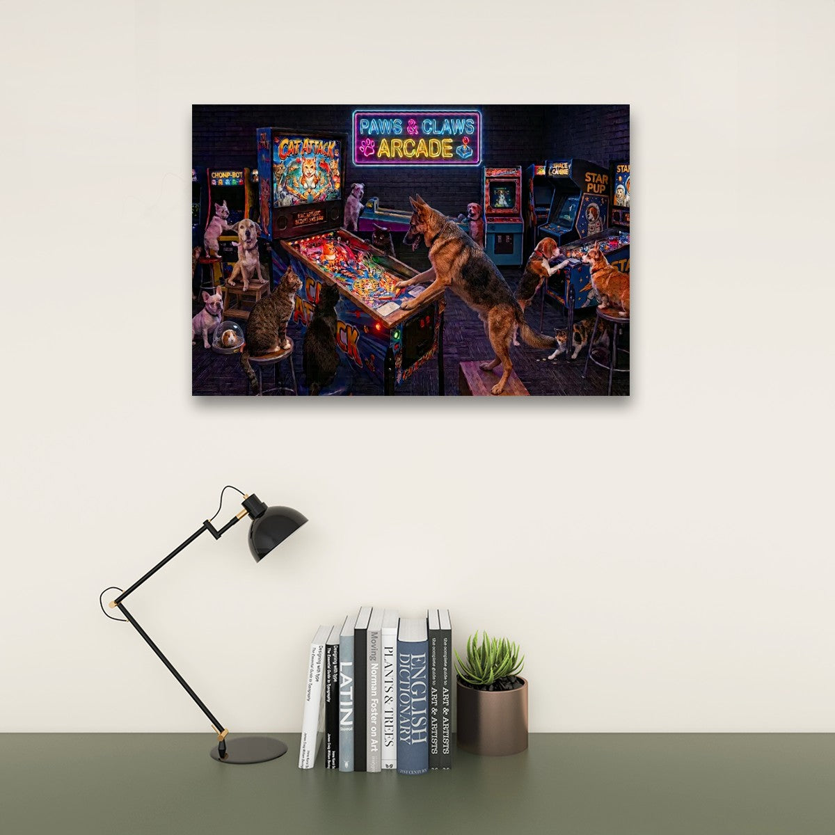 A framed wall art print depicting a retro arcade scene with dogs playing pinball machines, displayed above a desk with a black lamp, books, and a small potted succulent against a light beige wall.