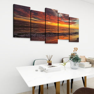 A five-panel canvas wall art set depicting a dramatic sunset over ocean waves, displayed on a white wall above a modern white dining table with chairs and decorative items.
