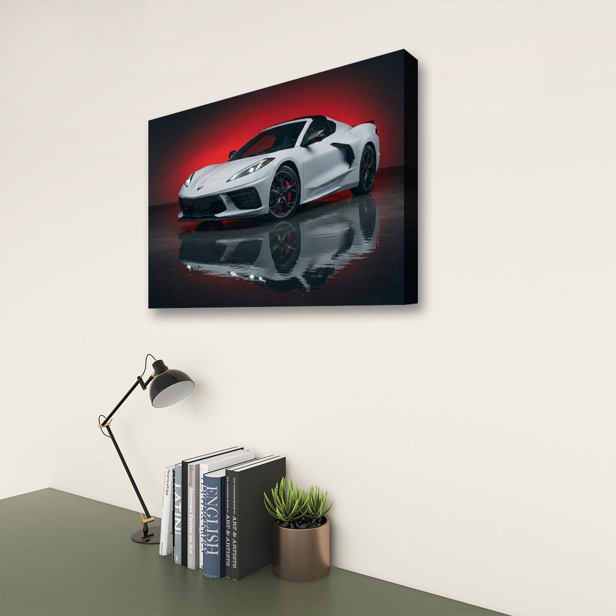 A rectangular wall art print showing a white Z8 Corvette sports car on a dark reflective surface with a red glow, displayed above a desk with a lamp, books, and a small plant in a room setting.
