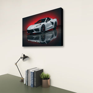 A rectangular wall art print showing a white Z8 Corvette sports car on a dark reflective surface with a red glow, displayed above a desk with a lamp, books, and a small plant in a room setting.
