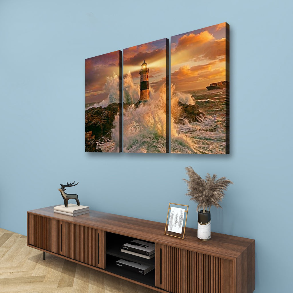 A three-panel canvas wall art set depicting a lighthouse on a rocky coast with dramatic orange and gold sunset sky, displayed above a wooden console against a light blue wall.