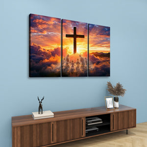 A three-panel canvas wall art set depicting a dramatic sunset sky with a large cross silhouette and glowing clouds, displayed above a wooden console table in a styled room.