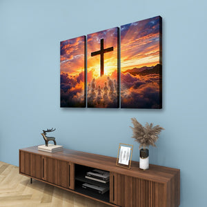 Three-panel canvas wall art showing a large black cross over a dramatic sunset sky with clouds and silhouetted figures, displayed above a wooden console in a styled room.
