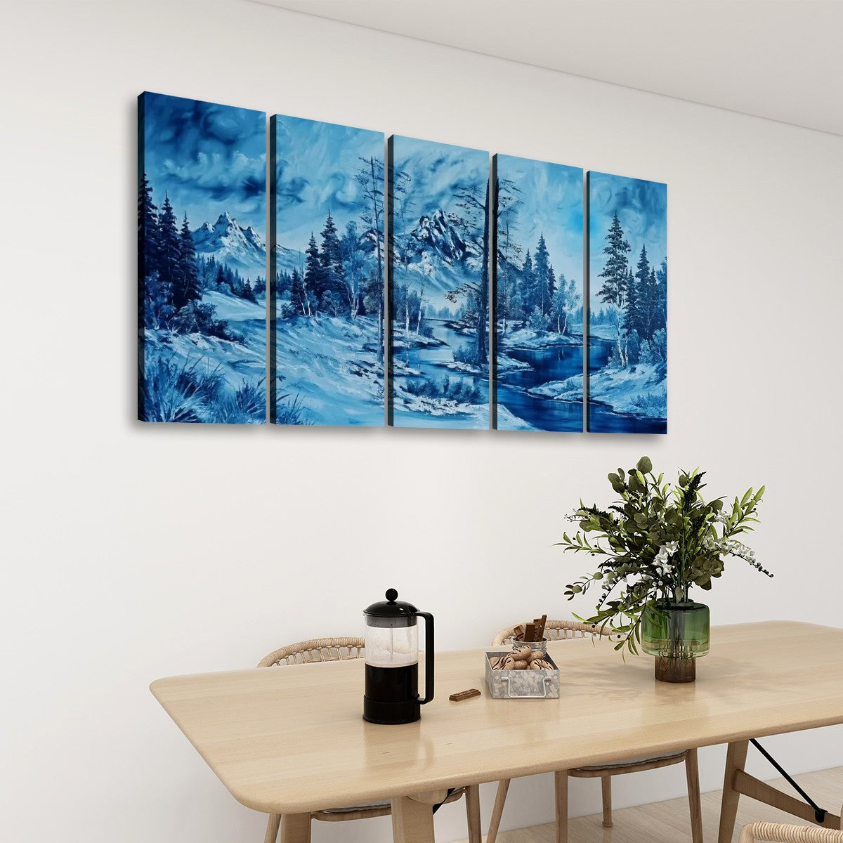 A five-panel canvas wall art set depicting a snowy mountain landscape in shades of blue, hung on a white wall above a light wood dining table with a coffee press and a potted plant.