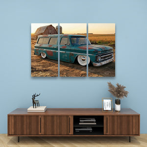 A three-panel canvas wall art print depicting a vintage teal 1966 Chevy Suburban Carryall parked in a rural field at sunset, displayed above a wooden console table against a light blue wall.