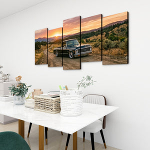A 5-panel canvas wall art set depicting a black vintage 1970 Chevy C10 Fleetside pickup truck on a dirt road at sunset, displayed on a white wall above a dining table in a modern room setting.