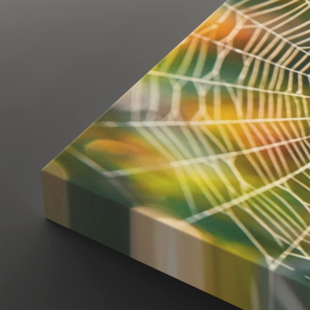 A close-up of a rectangular canvas or print showing a spiderweb pattern over a soft gradient background of green, yellow, and orange, photographed on a dark gray surface.