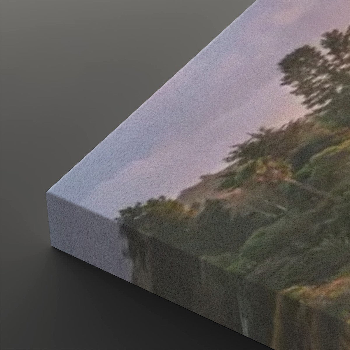 A close-up of a canvas print showing a landscape scene with trees, hills, and a soft pinkish sky, photographed at an angle on a dark gray background.