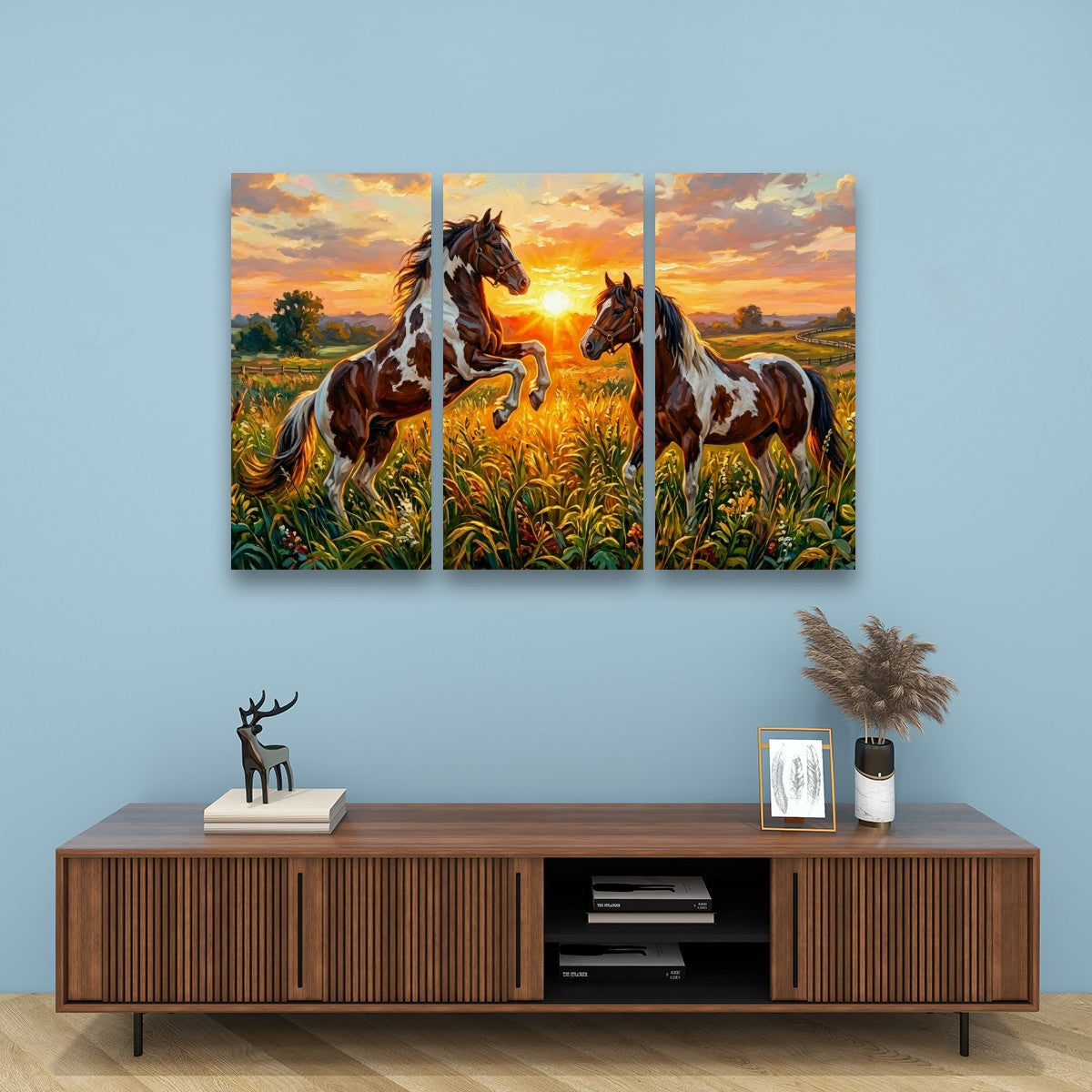 A three-panel canvas wall art set depicting two brown and white horses in a golden field at sunset, displayed on a light blue wall above a wooden console table with decorative items.