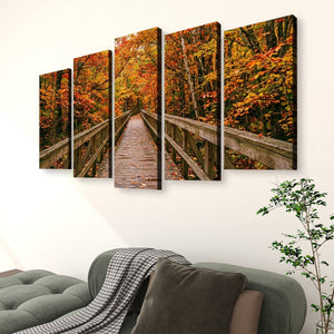 A five-panel canvas wall art set depicting a wooden boardwalk bridge through a dense autumn forest with orange, yellow, and red foliage, displayed on a white wall above a green sofa in a living room setting.
