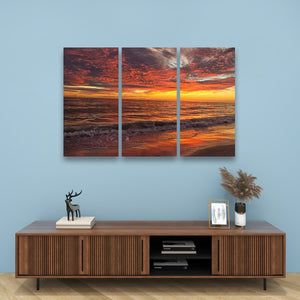 A three-panel canvas wall art set depicting a dramatic orange and red sunset over the ocean, displayed on a light blue wall above a wooden console table with decorative items.
