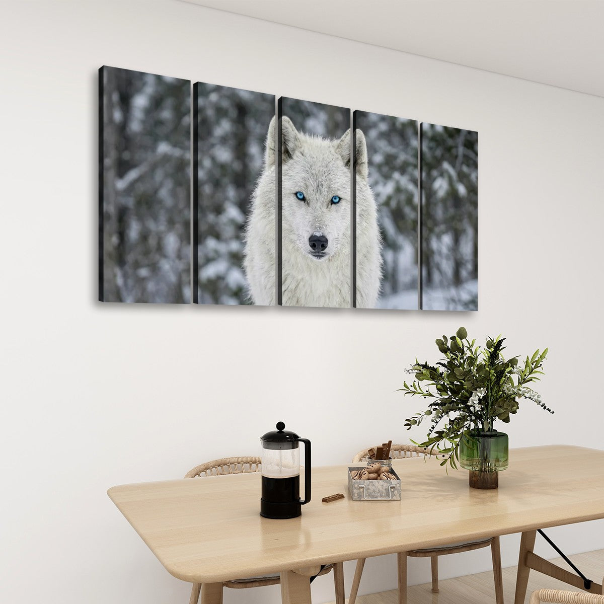 A five-panel canvas wall art set depicting a white wolf with bright blue eyes in a snowy forest, displayed on a white wall above a wooden dining table with chairs and a plant.