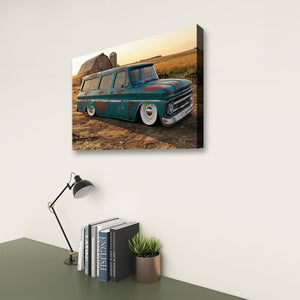 A rectangular wall art print showing a vintage teal 1966 Chevy Suburban Carryall parked outdoors near a barn and field, displayed above a desk with a lamp, books, and a small plant in a styled interior setting.