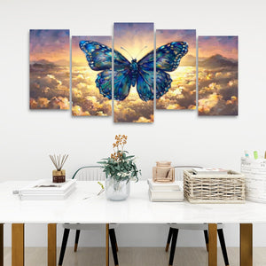 A five-panel canvas wall art set depicting a large blue butterfly over a golden sunset sky, displayed on a white wall above a styled dining table with books, a basket, and a vase.