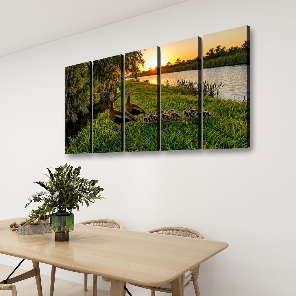 A four-panel canvas wall art set depicting a mother Black-bellied Whistling-Duck and her ducklings in a grassy field at sunset by a lake, displayed on a white wall above a wooden dining table with chairs.
