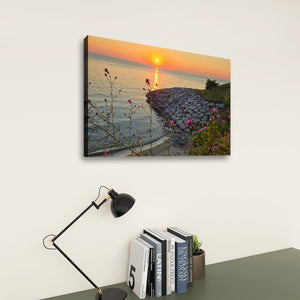 A rectangular wall art print showing a sunset over calm water with a rocky shoreline and flowers in the foreground, displayed on a light wall above a desk with a black lamp and books.