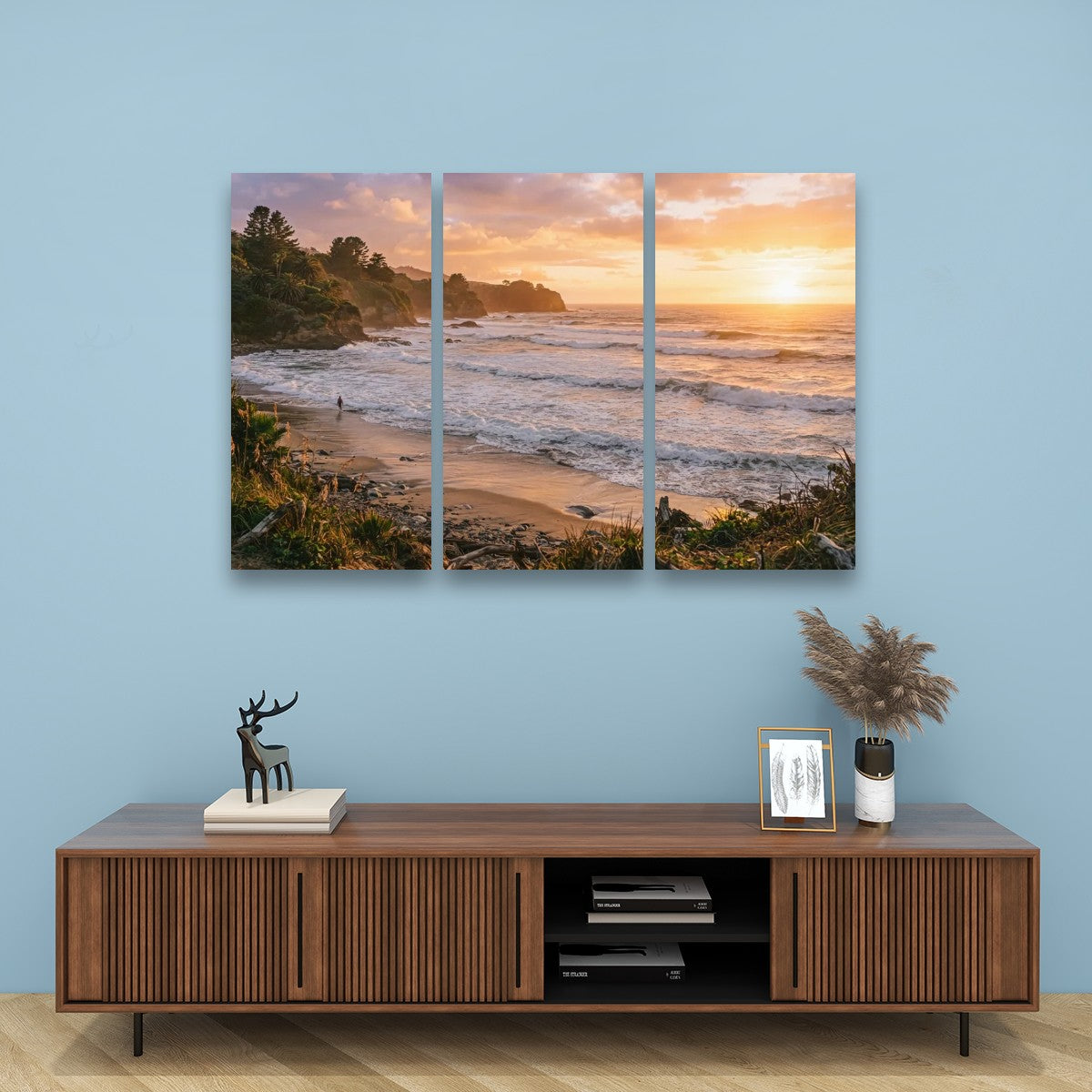 A three-panel canvas wall art set depicting a coastal beach sunset, displayed on a light blue wall above a wooden media console with decorative items.