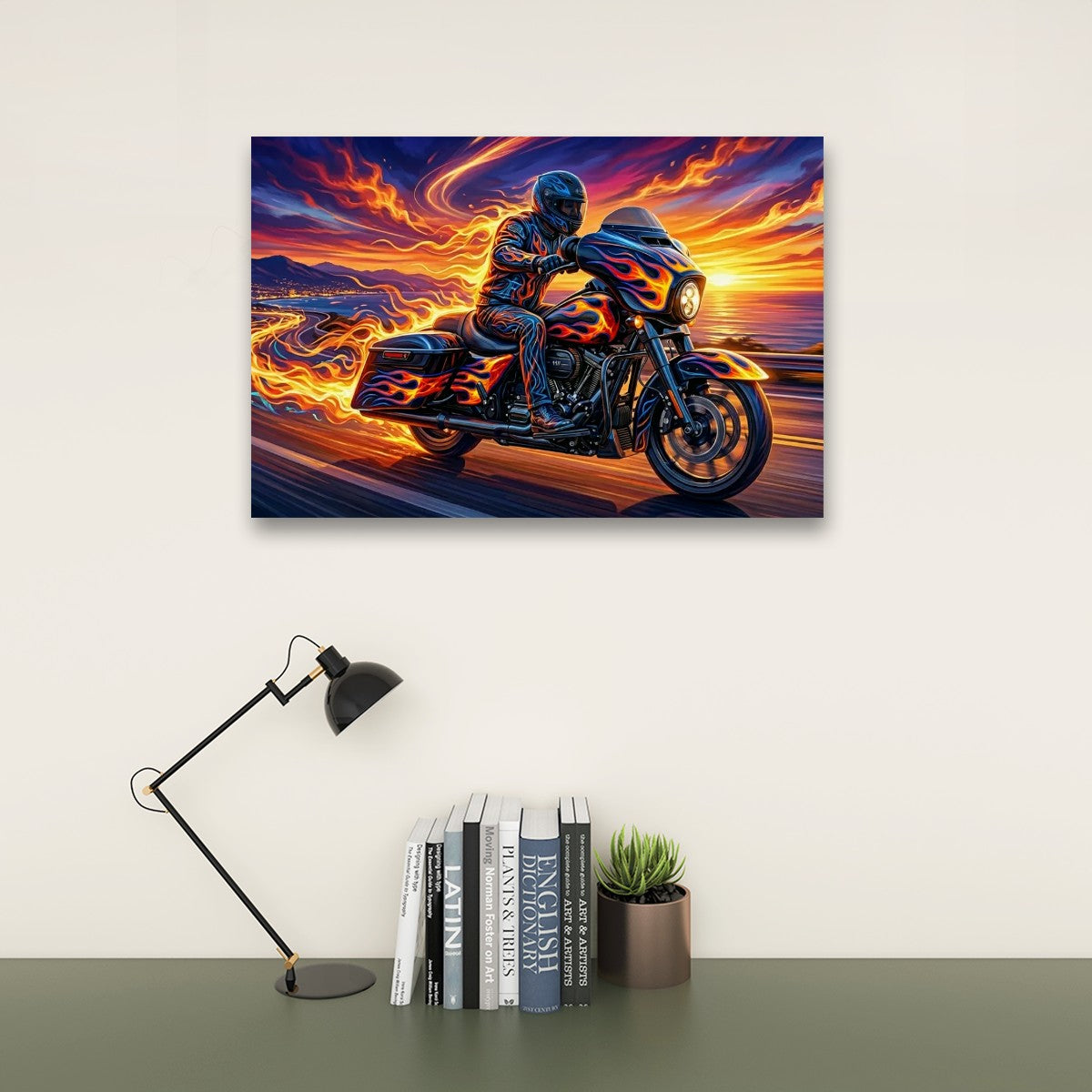 A rectangular wall art print showing a motorcycle rider on a flaming motorcycle against a dramatic sunset sky, displayed above a desk with a black lamp, books, and a small potted succulent.