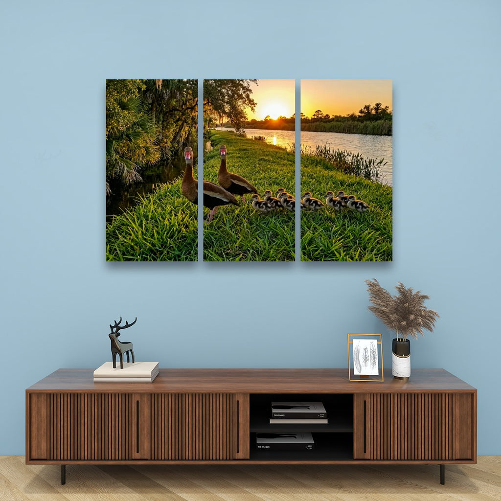 A three-panel canvas wall art print depicting a mother Black-bellied Whistling-Duck with her ducklings by a lake at sunset, displayed on a light blue wall above a wooden console table with decorative items.