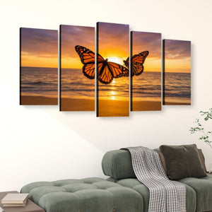 A five-panel canvas wall art set depicting a monarch butterfly flying over an ocean sunset, displayed on a white wall above a green sofa in a living room setting.