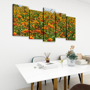 A five-panel canvas wall art set depicting a field of orange and yellow flowers with butterflies, displayed on a white wall above a modern white dining table with books and a vase.